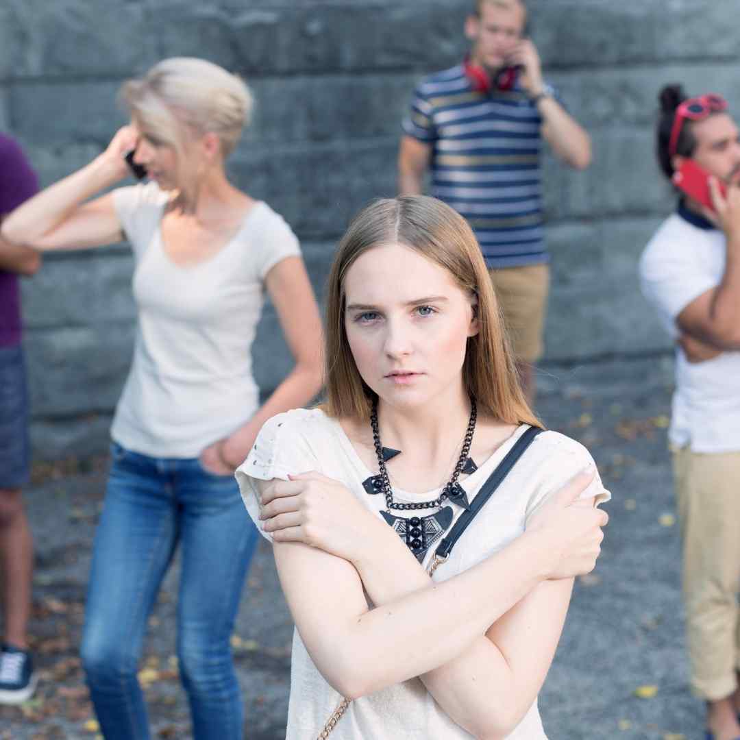 Eine Gruppe von Menschen, die aber mit sich selbst beschäftigt sind. Eine Person im Vordergrund verschränkt die Arme, blickt traurig in die Kamera