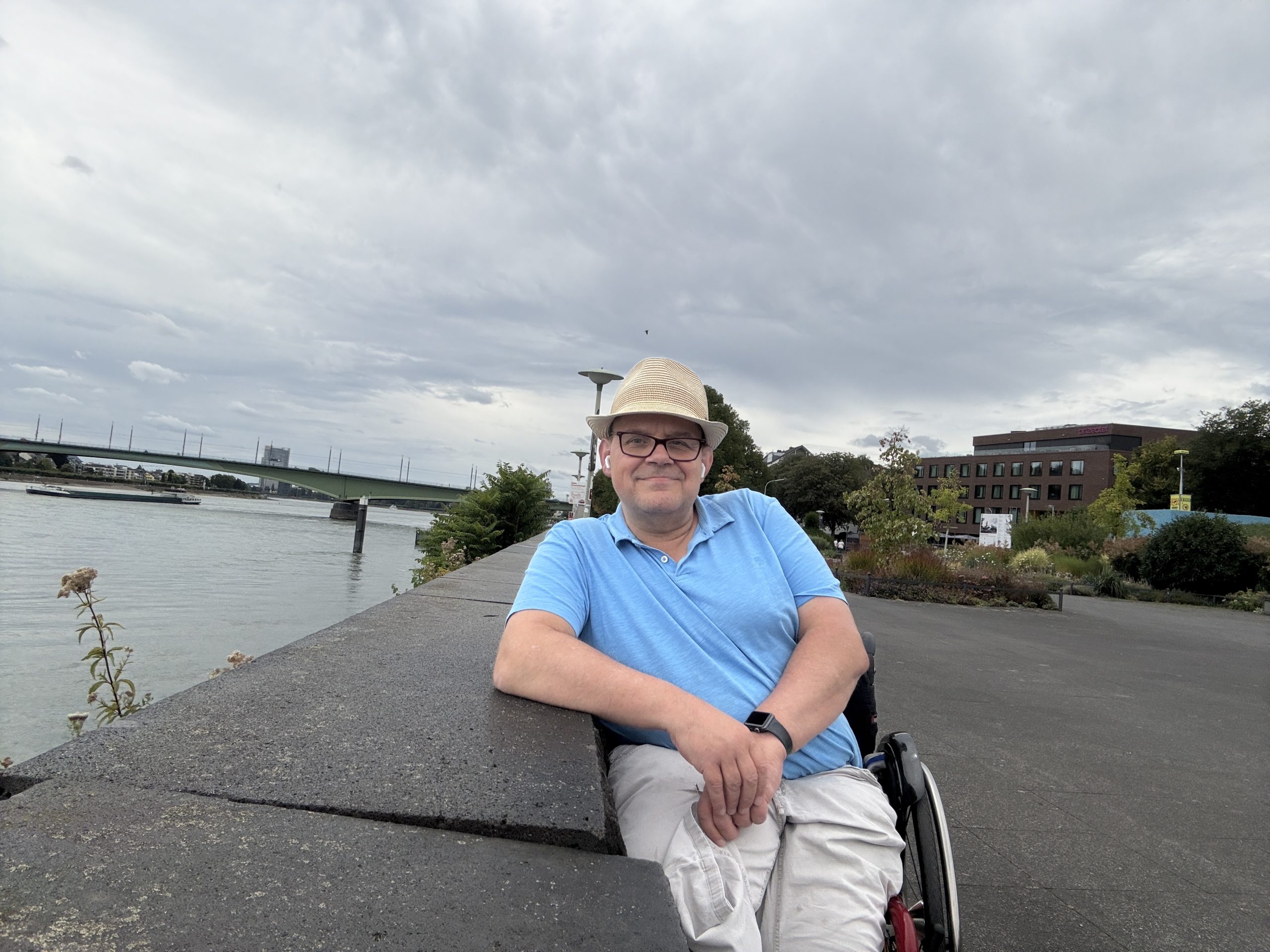 Ich im Sommer am Rheinufer mit hellblauen Poloshirt und heller Hose und einen Sommerhut auf dem Kopf